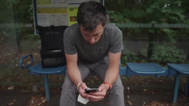 Mann Sitzt Bushaltestelle Wartet Auf Transport Und Surft Mit Smartphone — Stockvideo