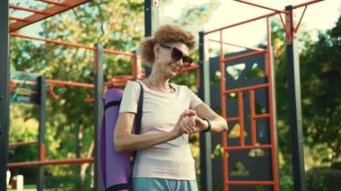 Yoga minderli yaşlı kadın spor salonunda duruyor ve spor saati kullanıyor. Yaşlı bir kadın yoga pedi tutuyor ve antrenmandan sonra kol saatini kontrol ediyor. Modern spor aletleri, fitness ekipmanları konsepti.