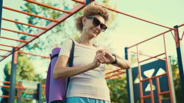 健美的老年妇女在外面上瑜伽课后使用健身跟踪器 现代体育用品 健身装备的概念 运动智能手表 退休时的健康生活方式 — 图库视频影像