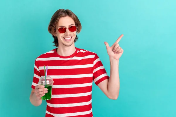 Фото молодого парня счастливая улыбка пить коктейли точка путешествия палец пустое пространство выбор объявлений изолированы на бирюзовом фоне — стоковое фото