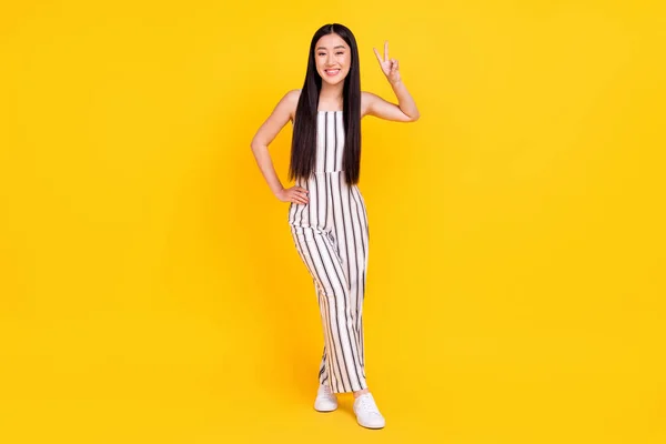 Plná délka tělo velikost fotografie dívka usmívá v pruhované celkově ukazuje v-znamení gesto izolované pulzující žlutá barva pozadí — Stock fotografie