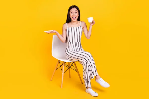 전체 몸길이의 사진 소녀웃으며 의자에 앉아 밝은 노란색 배경을 가진 카푸치노 컵을 놓고 미소짓고 있는 모습 — 스톡 사진