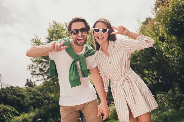 Portret fotograficzny młoda para uśmiechnięta latem idzie do parku w okularach przeciwsłonecznych pokazując gest v-sign — Zdjęcie stockowe