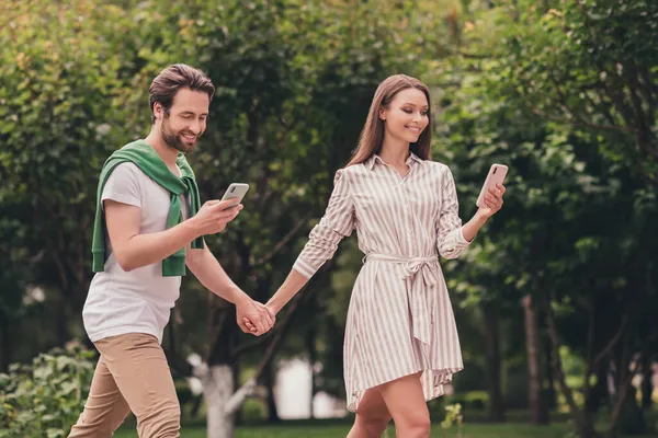 Foto portriat pareja joven sonriendo caminando en el parque verde tomados de la mano usando teléfonos inteligentes — Foto de Stock