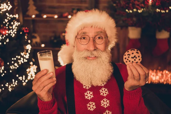 Foto de encantador homem maduro doce vestido Santa traje sorrindo segurando copo de leite saboroso biscoito dentro de casa quarto — Fotografia de Stock