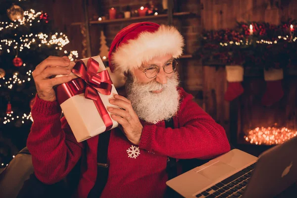 高级圣诞老人喜笑的照片拿着礼物盒在网上讲笔记本电脑在室内放暑假 — 图库照片