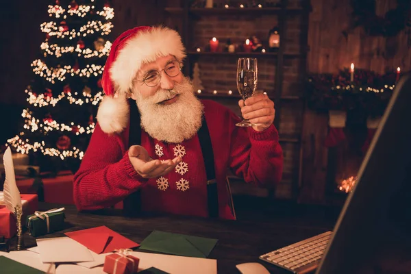 Фотопортрет веселый улыбающийся Санта пьет шампанское, разговаривая через веб-камеру в свитере с головными уборами — стоковое фото