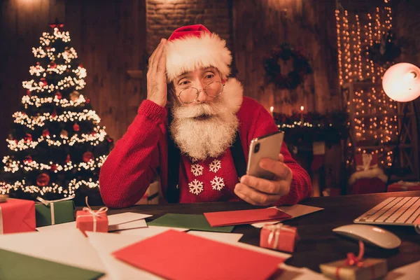 年迈的桑塔爪的照片惊奇的吃惊的手摸着头看浏览手机诺埃尔新年彩灯在室内 — 图库照片