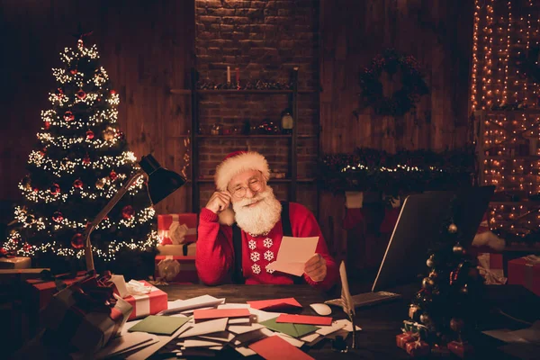 Санта Клаус улыбается на Рождество, сидя за столом и читая письма с пожеланиями в очках — стоковое фото