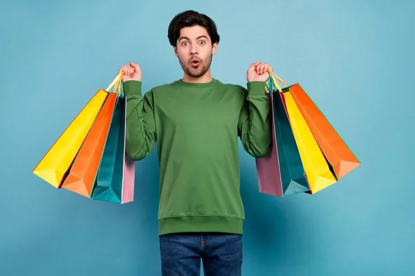Foto de joven sorprendido chico sorprendido en verde jersey shopaholic hold shop bolsas aisladas sobre fondo de color azul — Foto de Stock