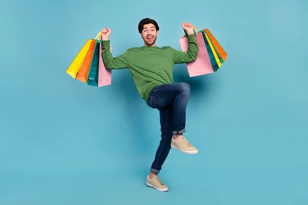 Foto em tamanho completo de jovem sorrindo homem levantar as mãos detém sacos de compras vencedor triunfo isolado no fundo de cor azul — Fotografia de Stock