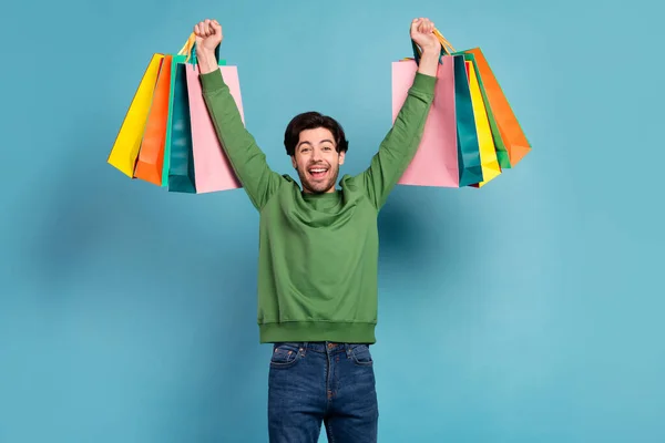 Foto von jungen lächelnden fröhlichen Mann heben die Hände yeah Hurra halten Einkaufstüten isoliert auf blauem Hintergrund — Stockfoto