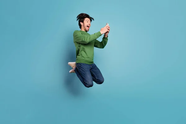 Photo de profil pleine grandeur côté de fou excité beau homme sautant en utilisant smartphone isolé sur fond de couleur bleue — Photo