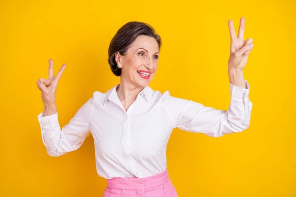 Foto de bonito engraçado idade mulher vestida camisa branca sorrindo mostrando v-sinal dançando olhando espaço vazio isolado cor amarela fundo — Fotografia de Stock