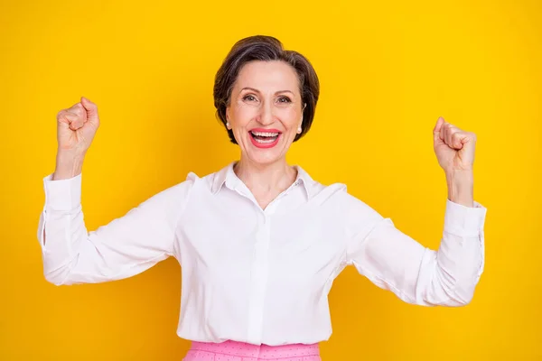 Foto de mulher adorável idade sorte vestida camisa branca levantando punhos sorrindo isolado cor amarela fundo — Fotografia de Stock