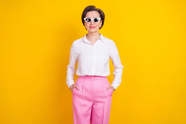 Fotografie rozkošný cool věk žena oblečená bílá košile tmavé brýle stojící ruce kapsy s úsměvem izolované žluté barvy pozadí — Stock fotografie