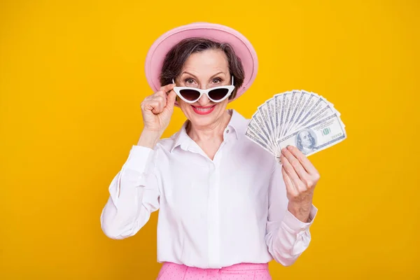Foto da mulher idosa feliz sorriso positivo segurar dólares dinheiro rico mão toque de vidro solar isolado sobre fundo de cor amarela — Fotografia de Stock