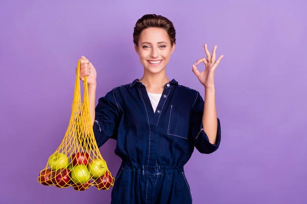 Foto di funky bruna giovane signora tenere frutta mostrano okey indossare jeans complessivamente isolato su sfondo di colore viola — Foto Stock