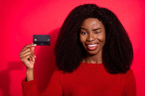 Foto de jovem afro menina piscar de olho feliz sorriso positivo segurar cartão de crédito bancário conselho de pagamento isolado sobre fundo de cor vermelha — Fotografia de Stock