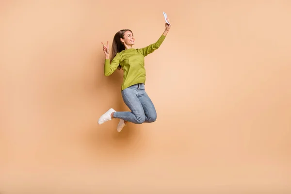 Perfil foto de menina salto segurar telefone fazer selfie mostrar v-sign desgaste verde pulôver jeans tênis isolado cor bege fundo — Fotografia de Stock