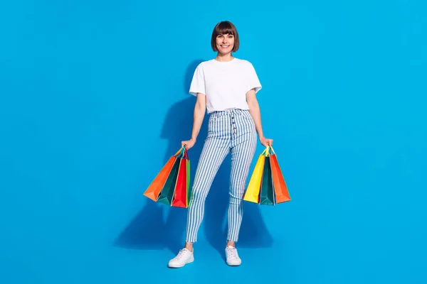 Fénykép csinos fényes fiatal nő öltözött alkalmi ruhák gazdaság bevásárlótáskák mosolygó elszigetelt kék háttér — Stock Fotó