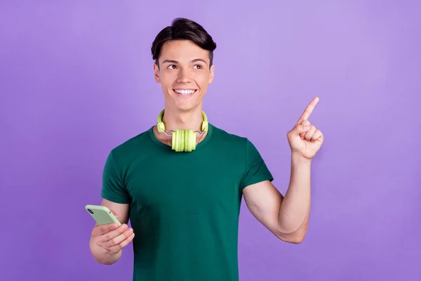 Zdjęcie marzycielski podekscytowany młody człowiek trzymać telefon punkt palec pusty przestrzeń słuchawki izolowane na fioletowy kolor tła — Zdjęcie stockowe
