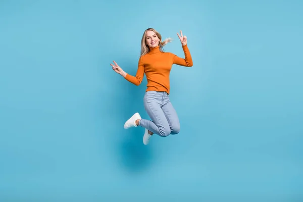 Fotografie lesklé sladké mladé ženy oblečené oranžový svetr s úsměvem skákání vysoko ukazuje V-znaky izolované modré barvy pozadí — Stock fotografie