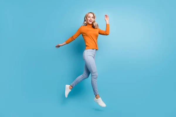 Foto de amigable joven bonita dama usar suéter naranja saltando alto brazo ondulante sonriendo aislado fondo de color azul —  Fotos de Stock