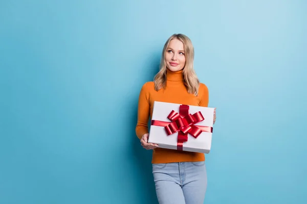 Fotografie okouzlující lesklá dáma nosit oranžový svetr držení dárek box hledá prázdný prostor s úsměvem izolované modré barvy pozadí — Stock fotografie