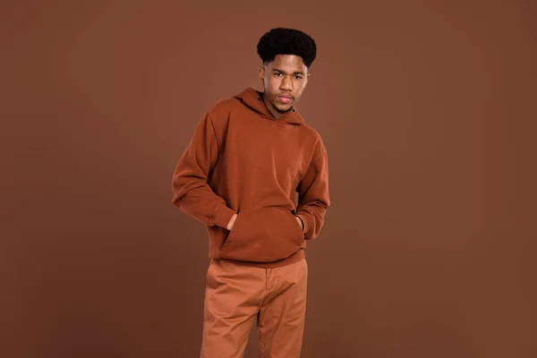 Foto do jovem afro bonito cara mãos no bolso desgaste sweatshirt sério isolado sobre cor marrom fundo — Fotografia de Stock