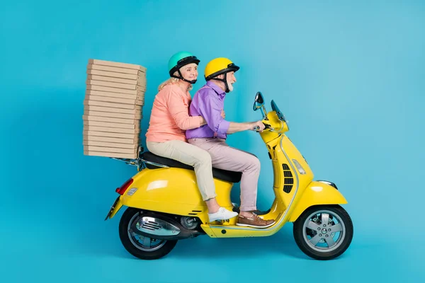 Portrait of two cheerful elderly retired pensioners riding moped delivering fastfood isolated over bright blue color background