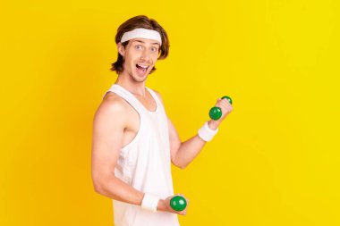Profile side view portrait of attractive cheerful guy lifting small light dumbbell isolated over bright yellow color background clipart
