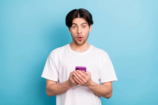 Foto van onder de indruk jonge brunet man houden telefoon dragen witte t-shirt geïsoleerd op blauwe kleur achtergrond — Stockfoto