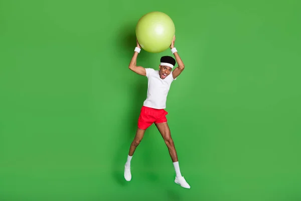 Foto de cuerpo entero de chocado divertido chico de piel oscura vestido traje deportivo lanzando bola en forma saltando aislado color verde fondo —  Fotos de Stock