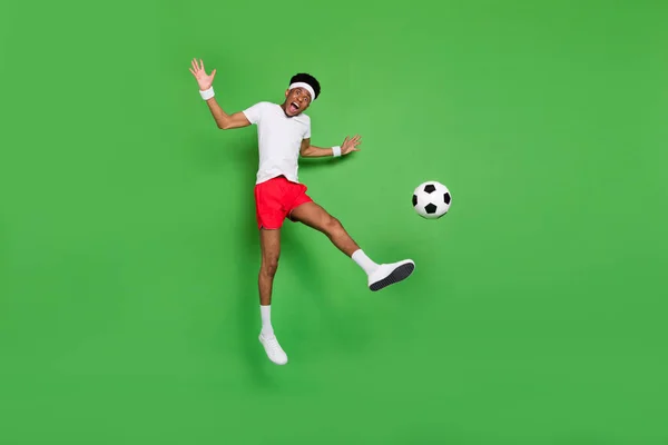 Полная длина фото сильной возбужденной темной кожи парень одет спортивный костюм прыжки играть футбол изолированный зеленый цвет фона — стоковое фото