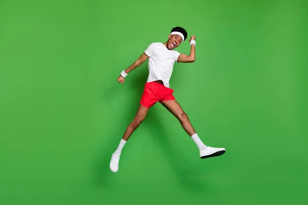 Lunghezza completa foto di impressionato eccitato pelle scura uomo indossare bianco t-shirt alzando pugni saltando alto isolato colore verde sfondo — Foto Stock