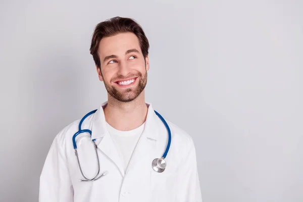 Foto van professionele positieve jonge doc man gekleed witte jas glimlachen op zoek lege ruimte geïsoleerde grijze kleur achtergrond — Stockfoto