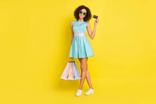 Foto de cuerpo completo del divertido peinado rizado mujer joven sostiene bolsos tarjeta de desgaste azul vestido gafas aisladas en el fondo de color amarillo —  Fotos de Stock