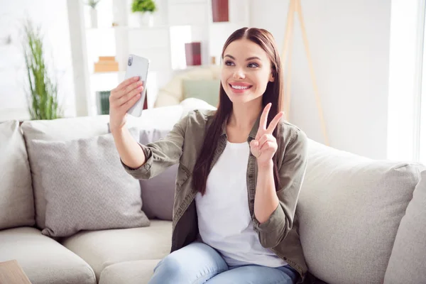 Yeşil tişörtlü tatlı bir kadının fotoğrafı kanepeye oturmuş selfie yapıştırıyor modern aletler ev içinde v-işareti gösteriyor. — Stok fotoğraf