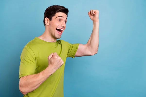 Foto de dulce afortunado joven usar camiseta verde sonriendo puños levantándose buscando espacio vacío aislado color azul fondo —  Fotos de Stock