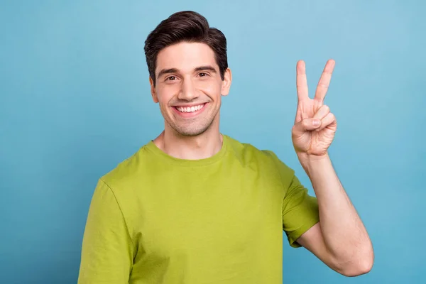 Фото гарного настрою солодкий молодий хлопець одягнений в зелену футболку, що показує v-знак усміхнений ізольований синій колір фону — стокове фото