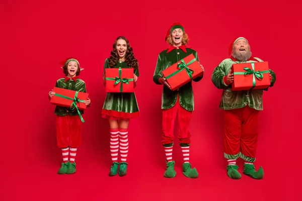 Volledige lengte foto van vrolijke santa helper houden geschenk doos genieten van kerst traditie slijtage hoed geïsoleerd op rode kleur achtergrond — Stockfoto