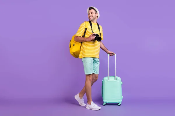 Pełne ciało zdjęcie brunet szczęśliwy pozytywny człowiek spacer bagaż bagaż aparat casual ubrania kapelusz izolowany na fioletowy kolor tło — Zdjęcie stockowe