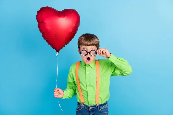 Фото изумленного шокированного мальчика, держащегося за руки шарик в форме сердца носить очки изолированы на синем фоне цвета — стоковое фото