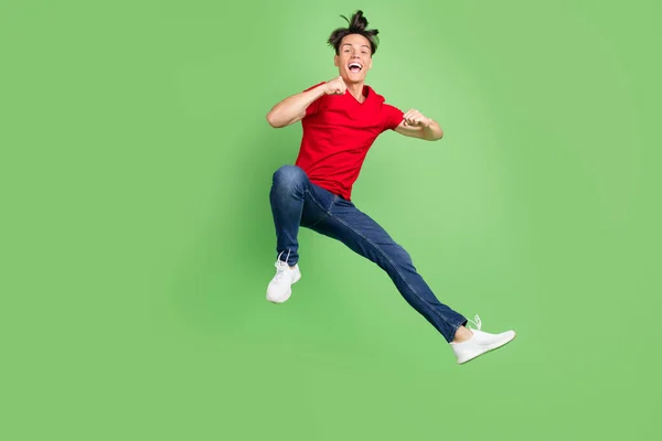 Foto van grappige opgewonden jonge man gekleed rood t-shirt springen hoog stijgende vuisten glimlachen geïsoleerde groene kleur achtergrond — Stockfoto