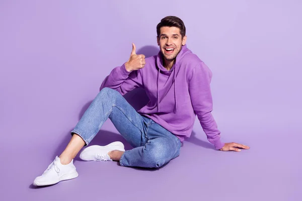 Full size corpo formato foto uomo sorridente mostrando come segno seduto sul pavimento isolato colore viola pastello sfondo — Foto Stock