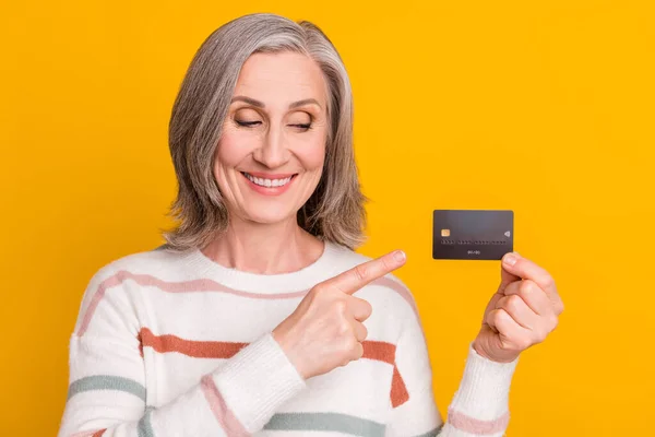 Foto de muito doce senhora pensioner desgaste listrado pulôver olhando apontando cartão de crédito sorrindo isolado cor amarela fundo — Fotografia de Stock