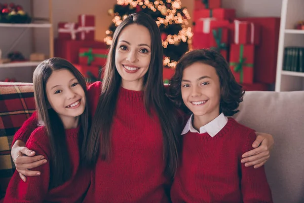 Фото мечтательной сладкой мамы двое детей, одетых в красные свитера, обнимающие улыбающиеся, празднующие Новый год в помещении — стоковое фото