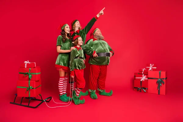 Volledige grootte foto van santa helpers mensen wijzen kijken lege ruimte met slee giftboxen dragen nieuwjaar kostuums geïsoleerd op rode achtergrond — Stockfoto