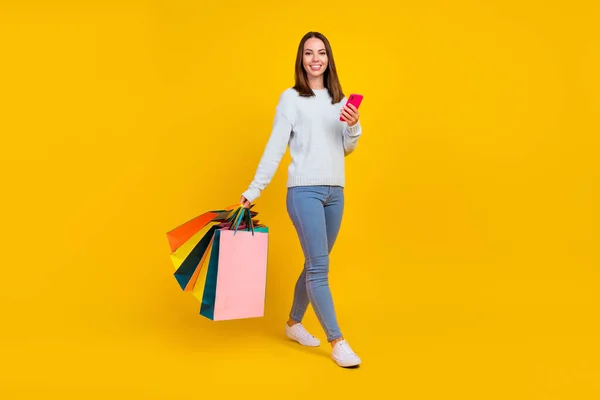 Foto corpo pieno di moda felice giovane donna dai capelli castani tenere telefono camminare borsa shopper isolato su sfondo di colore giallo — Foto Stock